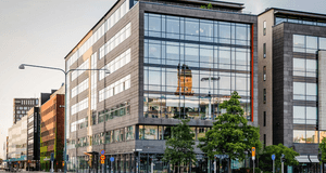 Exteriör av en modern kontorsbyggnad i glas och mörk sten vid Hjälmarekajen. I den stora glasfasaden reflekteras ett äldre torn, och framför byggnaden syns gröna träd, gatubelysning och en tom väg i mjukt dagsljus.
