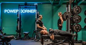 En kvinna gör hantelpress på en träningsbänk i ett gym med turkosa väggar och neonskylten "POWER CORNER".