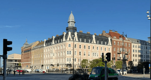 Exteriörbild i grodperspektiv över ett stadslandskap med en klassisk hörnbystnad med torn, omgiven av trafik och korsning under en klarblå sky.