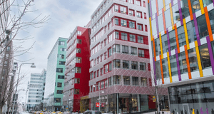Gatuliv i ett modernt kontorskvarter med färgstarka fasader i Stockholm. I mitten syns en röd byggnad med mönstrat metallnät över glaspartier, omgiven av höghus med gröna, vita och flerfärgade fasader. Flera bilar, inklusive gula taxibilar, kör längs gata
