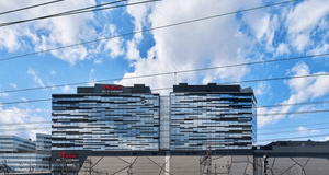 Exteriörbild av Westfield Mall of Scandinavia i Solna, Stockholm, sedd från spårområdet. Fasaden består av två svarta och glasbeklädda kontorsbyggnader med ett abstrakt, mosaikliknande mönster i blått, vitt och svart. I förgrunden syns flera tågledningar 