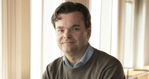 Ett porträtt av en man med mörkt, vågigt hår, klädd i en ljusblå skjorta under en brun stickad tröja. Han tittar direkt in i kameran med ett neutralt uttryck. Bakgrunden är ljus och mjukt suddig med vertikala linjer från fönsterpartier som släpper in natu