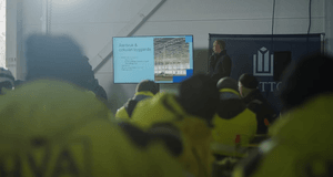 En föreläsare håller en presentation om återbruk och cirkulärt byggande inför en publik i gula varselkläder i en industriell lokal.