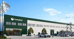 Digital rendering (3D-bild) av en stor, modern industrilokal eller ett lagerhus i vitt och mörkgrönt. På fasaden står logotypen "Coloplast". Framför byggnaden finns en parkeringsplats med flera bilar, inklusive laddningsstationer, och flaggor med logotype