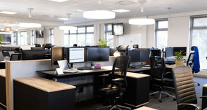 Ett ljust och modernt kontorslandskap med tomma arbetsplatser. Skrivborden är utrustade med dubbla bildskärmar, bärbara datorer och ergonomiska kontorsstolar. Stora runda taklampor ger ett mjukt ljus över lokalen.