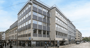 En hörnfastighet i flera våningar med grå och beige fasad i en stadsmiljö. Byggnaden ligger i korsningen mellan en gågata med flanörer och en bilväg, med butiksfönster på bottenvåningen.