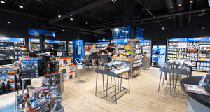 Detta är en interiörbild av en modern elektronikbutik eller butik som säljer teknik och relaterade produkter. Lokalen har en mörk inredning med svarta väggar och tak, där synliga ventilationsrör och kablar bidrar till en industriell känsla. Golvet är ljus