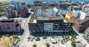 Flygfoto över ett torg och stadsområde med en stor mörk fastighet utrustad med solpaneler och växttak vid havet.