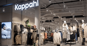 Interiörbild av en modern Kappahl-butik. Bilden visar entrén med en upplyst logotyp och en stor digital skärm med reklam. Inuti butiken hänger kläder i höstfärger på ställ, och flera skyltdockor visar upp stickade plagg och ytterkläder. En kvinna med röd 