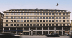 En bred fasadbild av en stor kontorsbyggnad i flera våningar vid en trafikerad stadsgata. I gatuplan syns butiker och verksamheter som Visita, Fabrique och ett apotek. Suddiga bilar i rörelse passerar framför byggnaden under en klar himmel.