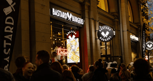 Kvällsbild på en Bastard Burgers-restaurang med upplysta skyltar och en folksamling som väntar utanför entrén.