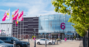 Väla köpcentrums entré nummer 6 med en stor rundad glasfasad, flaggor som vajar i vinden och parkerade bilar i förgrunden.