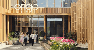 En modern byggnad med namnet "Origo" ovanför entrén. Fasaden är klädd i ljusa träribbor och stora glaspartier. I förgrunden syns en stenlagd gångväg kantad av blommande rosa och vita tulpaner, och en grupp människor är på väg in i byggnaden.