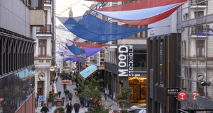 En vy över en tät stadsgata/gågata i skymningen eller på kvällen, dominerad av byggnader med äldre arkitektur och moderna inslag. Gatan är dekorerad med stora, spända tygsegel i blåa, röda och lila nyanser som hänger över gångvägen. Längre fram i bilden s