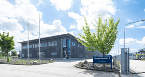 Exteriörbild av en modern, mörkgrå industribyggnad i två våningar, med en entré i glas. Framför byggnaden syns en asfalterad uppfart och en blå skylt som anger adressen "Olsgårdsgatan 3" och namnet "Wihlborgs". Omgivningen har grusade ytor och välklippta 