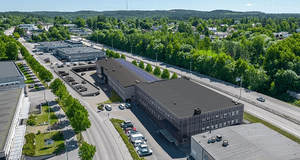 Flygfoto över ett industriområde med kontors- och lagerbyggnader omgivna av vägar, parkerade bilar och grönskande skogsområden i bakgrunden under en blå himmel.