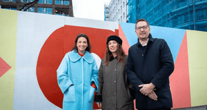 Tre personer, två kvinnor och en man, står framför ett byggplank målat med stora, färgglada geometriska former i rött, orange, rosa och gult. Kvinnan till vänster bär en ljusblå kappa, kvinnan i mitten bär en mörk kappa och basker, och mannen till höger b