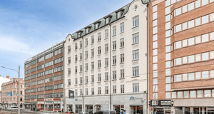 En ljusgul, sekelskiftesliknande flervåningsbyggnad placerad mellan två mer moderna kontorshus i tegel och glas längs en stadsilag.