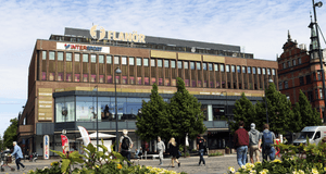 Gallerian Flanör i Gävle sett från Stortorget en solig dag. I förgrunden syns gula blommor och människor som rör sig på torget framför den stora tegelbyggnaden, som även har skyltar för Intersport.