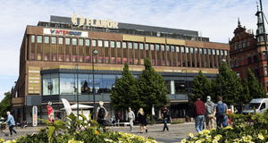 Exteriör av gallerian Flanör i Gävle sett från Stortorget. Den bruna tegelbyggnaden har stora glaspartier på gatuplan, skyltar för butiker som Intersport och en takskylt med namnet Flanör. Människor promenerar på det stenlagda torget framför gula blommor 