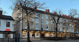 Bild tagen under kvällstid av ett bostadshus med tre våningar i ljusgul puts. På gatuplanet finns flera butiker under ett utskjutande tak med inbyggd belysning. Flera kala träd står framför huset, varav ett är dekorerat med julbelysning