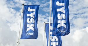 Tre blå flaggor med Jysk-logotypen i vitt som svajar i vinden mot en blå himmel med vita moln.