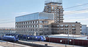 Blå pendeltåg vid en tågstation framför en modern tegelbyggnad med stora glaspartier och en hög huskropp.