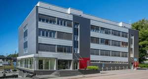 Modern kontorsbyggnad i fyra våningar med grå fasadpaneler, röda accentdetaljer och entrétrappa mot gatan under en klarblå sky.