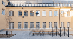 En stenlagd innergård framför en klassisk, ljusgul byggnad med regelbundna fönsterrader, bänkar och svarta metallskärmar.
