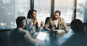 Fyra affärsklädda personer sitter i ett möte runt ett bord i ett ljust konferensrum med stora fönster. En kvinna i mitten talar engagerat och gestikulerar, medan de övriga lyssnar uppmärksamt. På bordet syns bärbara datorer och surfplattor.