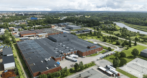 En flygbild över ett industriområde med stora lagerbyggnader i rött tegel och plåt. Flera lastbilar står parkerade vid lastkajer och på en intilliggande parkering. I bakgrunden syns ett grönskande landskap med en ringlande å, skogspartier och ett bostadso