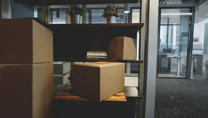 Flyttkartonger i papp staplade på möbler i en modern kontorsmiljö. I bakgrunden syns hyllor med böcker och krukväxter, samt glasväggar som vette mot andra delar av kontoret och fönster med utsikt över stadens höghus.