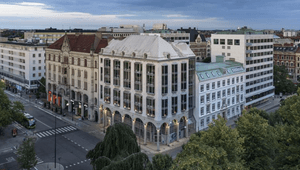 Flygfoto i fågelperspektiv över ett stadskvarter med en ljus hörnfastighet med valvbågar i gatuplan under en molnig skymningshimmel
