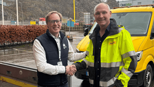 Två män skakar hand utomhus framför ett industriområde och ett gult fordon. Mannen till vänster bär glasögon, en vit skjorta och en mörkblå väst. Mannen till höger bär en gul och grå varseljacka med reflexer.