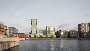 Ett stadslandskap vid vattnet med moderna bostads- och kontorshus i olika färger och höjder. I mitten syns en ljusgrön byggnad med en högre tornsektion och en bredare lägre del. Till vänster finns en röd gångbro över vattnet, och runtomkring står andra hu