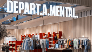 En modern butik med namnet "DEPART.A.MENTAL" i stora vita lysande bokstäver i taket. Inuti syns klädställ med skjortor, klänningar och tröjor i olika färger, samt hyllor med hudvårdsprodukter och accessoarer. Inredningen är ljus med röda hyllmoduler, träd
