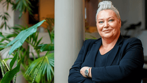 Ett porträtt av en leende kvinna med kort blont hår, iklädd en svart kavaj, som lutar sig mot en pelare med gröna växter i bakgrunden.