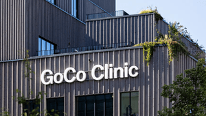Exteriör av GoCo Clinic. Den moderna byggnaden har en fasad av lodräta träribbor och stora vita bokstäver som bildar namnet. På takterrasserna syns gröna växter och hängande klätterväxter mot en klarblå himmel.