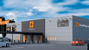 Byggnaden har en stram, industriell design med grå fasad och en framträdande mörkgrå sektion i mitten där den orangea Snabbgross-logotypen är placerad. Till höger om logotypen syns butikens öppettider listade, bland annat med tidiga öppettider på vardagar