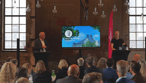 En presentation pågår i en industrilokal med stora fönster och många hängande glödlampor i taket. Två män står vid varsin talarpod och talar inför en sittande publik. Mellan dem syns en stor skärm med en presentation som visar ett porträttfoto, namnet And