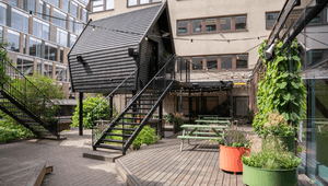 En modern takterrass med en unik svart trästuga upphöjd på stolpar. Terrassen har trädäck, sittplatser vid gröna bänkbord, stora färgstarka krukor med växter och ljusslingor som hänger över ytan. I bakgrunden syns urbana kontorsbyggnader.