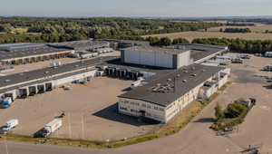 Flygfoto i fågelperspektiv över ett stort logistik- och lagerkomplex med lastkajer, lastbilar och omgärdande åkermark.
