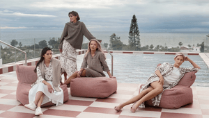 Fyra kvinnor poserar i stilrena kläder i jordnära toner på en takterrass med pool. De sitter och står vid rosa sittpuffar på ett schackrutigt golv, med havet och en palm i bakgrunden under en molnig himmel.
