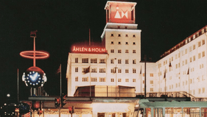 Klassiskt nattfoto på Åhlén & Holms byggnad med upplysta skyltar och ett torn. I förgrunden syns en ljusinstallation och en spårvagn.