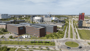 En vy från ovan över ett modernt kontorsområde med varierad arkitektur och mycket grönska. I förgrunden löper en väg där en grön spårvagn passerar en rondell. Till vänster syns stora bruna tegelbyggnader med solpaneler på taken, och i bakgrunden sticker e