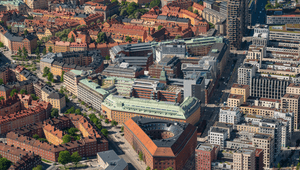 En flygbild över en tätbebyggd stadsmiljö med en unik triangelformad tegelbyggnad i centrum, omgiven av klassiska husrader och moderna höghus under en klarblå himmel.