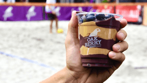 En hand håller upp en genomskinlig bägare med en açaí-bowl från Oakberry. Innehållet är varvat med lila açaí och jordnötssmör, toppat med blåbär. I bakgrunden syns en sandplan där personer spelar sport.