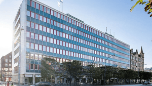 En exteriörbild tagen på gatunivå visar en stor, modern kontorsbyggnad i flera våningar med en fasad dominerad av långa rader med fönster. Fönstren har blågröna glaspartier och kontrasterande röda fönsterkarmar. Byggnadens bottenplan har skyltfönster och