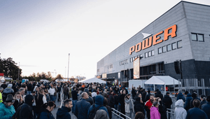 En stor folkmassa köar utanför en stor Power-elektronikbutik under dagsljus. Människor står tätt i fållor avgränsade med metallstängsel framför den grå butiksbyggnaden med den orangea Power-logotypen.