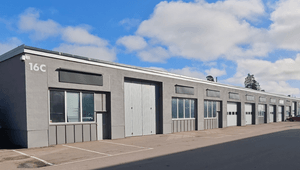 En lång, grå industrifastighet i en våning med flera sektioner under en blå himmel med lätta moln. Byggnaden har stora fönsterpartier, industriportar och en markerad husnummering "16C" på hörnet. Framför byggnaden finns en asfalterad yta med parkeringsmar